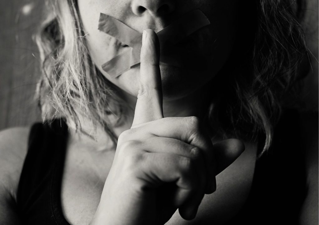 Woman holding finger to lips, mouth has tape across it